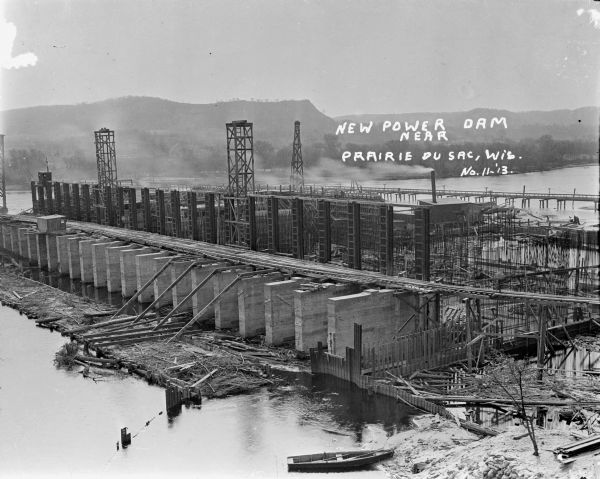 Elevated view of a power dam on the Wisconsin River under construction. There is a rowboat in the foreground. On the opposite shoreline are trees and bluffs.