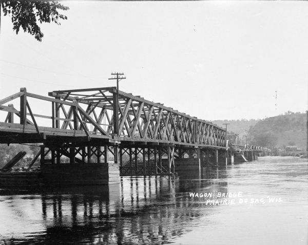 View across water towards a wooden wagon bridge over the Wisconsin River. A farm and tree-covered hills are on the opposite shoreline.