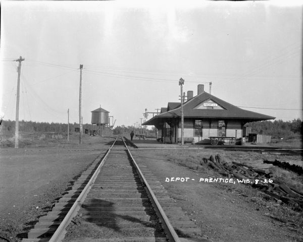 View down center of railroad tracks towards the Prentice Depot and railroad crossing. A man is walking on the platform of the depot. On the left in the background is a water tower.