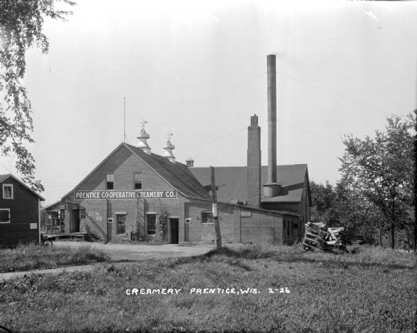 View across yard of the co-operative Creamery Company. There are two large vents on the roof of the building that have weather vanes decorated with cows.
