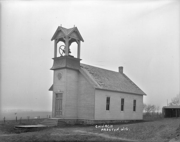Wooden church with a bell tower and a stable in the back on the right. Fences and fields are in the background.
