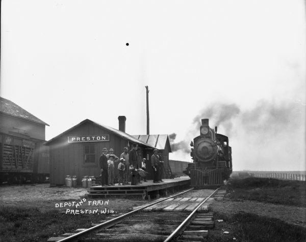 View along railroad tracks towards a group of men and boys standing on a platform in front of the Preston Depot while a locomotive arrives. In the background freight cars are sitting on another set of tracks, and a large building is in the background.