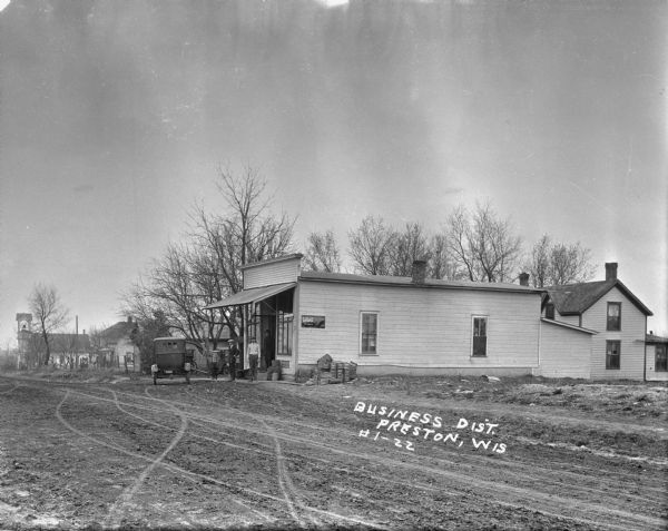 View across unpaved road of the Preston Business District. Three men and a young child stand in front of what may to be a general store, with large show windows. Automobiles are parked around the store, and dwellings and other buildings are in the background. There is a church with a bell tower further down the road.