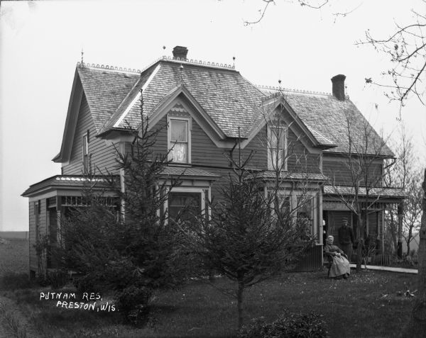 View of Putnam residence and post office, with Mrs. Putnam sitting in a chair on the lawn, and Mr. Putnam (with a pipe in his mouth) standing behind her on the walk in front of the porch. Pine trees are in the yard.