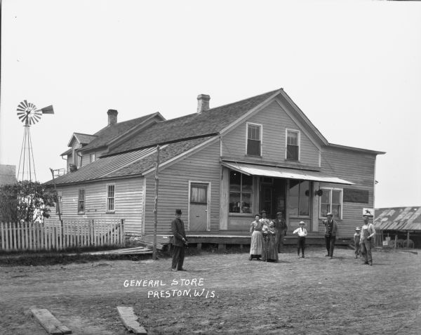View across street of the General Store. A group of five men, two women, and three children are standing in front. with a windmill is in the back and a stable on the side. A white picket fence on the left.