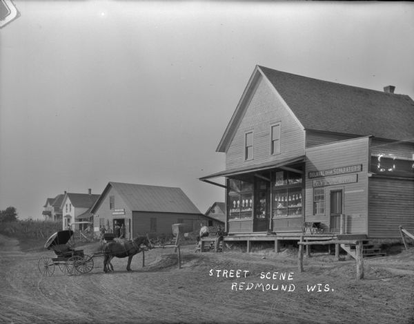View across street of a general store on the right, and another business on the left. Two men and a dog pose in front of the general store. A horse and buggy stand in the road. Three men and a young boy stand in the open doorway of the building on the left below a sign that says "Imperial Drills" and "LaCrosse." Further down the street are houses and a field.