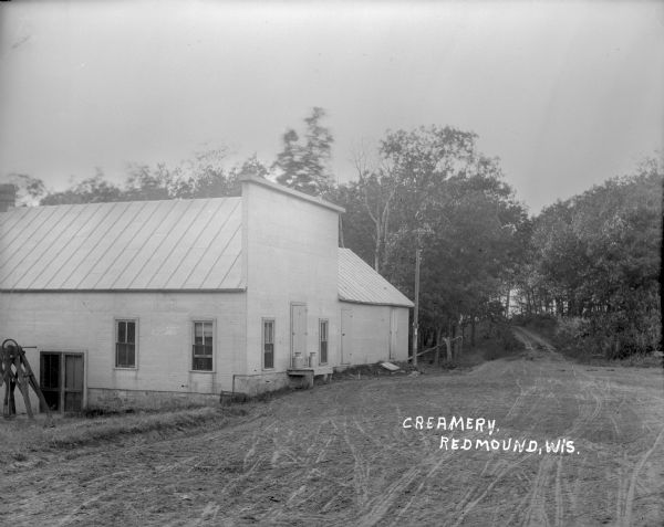 View from dirt road of a creamery. Two milk cans stand on a small loading dock. The road continues on through woods in the background.