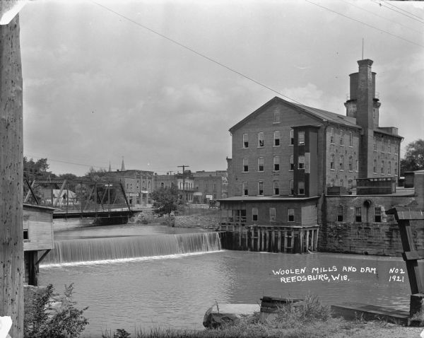 View from shoreline over the Baraboo River towards a dam, with a mill on the opposite shoreline. A bridge and a business district are in the background.