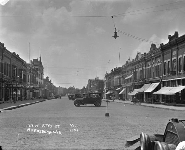View down Main Street. A millinery, meat market, bakery, hotel and shoe store are on the left. An osteopath, a hardware store, a barber shop, a small restaurant and a department store are on the right. Automobiles are parked in the center of the street. Pedestrians are on the sidewalks.