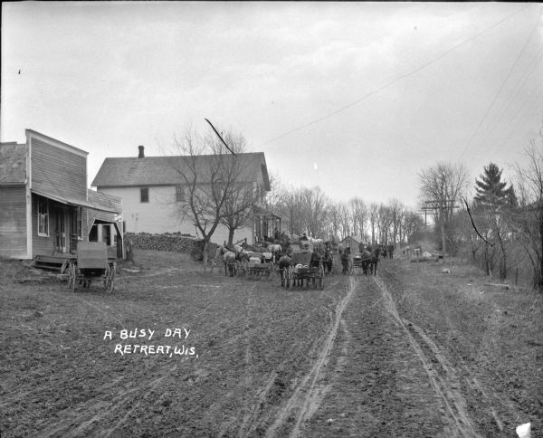 View down dirt road of horses and delivery wagons parked near wooden buildings on the left. A group of people are assembled, some on the porch of a large building, and some in the road.