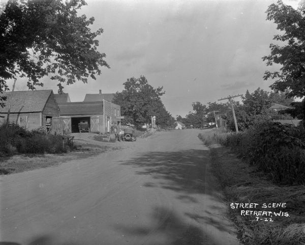 View down dirt road towards a garage and other wooden buildings on the left. A tire shop back on the right. Two men are standing in front of the garage near an automobile parked at the curb. Another automobile can be seen through the open door of the garage. Further down the road a horse and buggy are parked in front of another storefront.