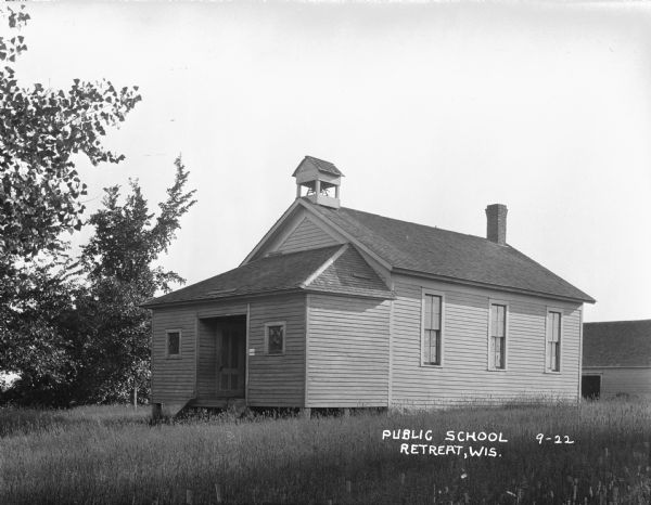 View of the public school in a rural area. The front door is inset, and two small square windows flank the entrance. A small structure with a roof houses a bell. Behind the school is a wooden structure with a large open door. Tall grass and trees are in the yard.