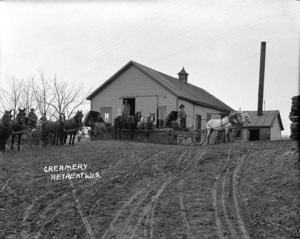 View across dirt road of a group of men posing with horses and wagons. One man stand on the loading dock. There is a tall smokestack on a section of the building in the back. Firewood is stacked on the right.