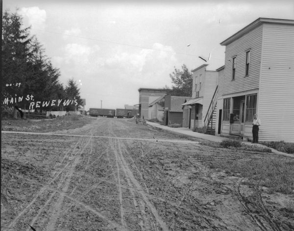 View down unpaved Main Street towards a train passing by in the background. There is a row of buildings on the right and a fenced in lot on the left. A wooden cart is parked in the road. A man stands on the sidewalk at the corner of the building on the right. Near the train is the Rewey Hotel on the left, and a livery on the right.