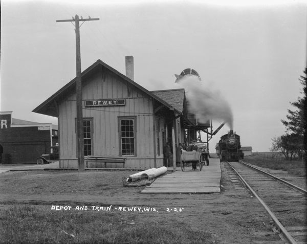 View along railroad tracks towards the Rewey railroad station. A locomotive spewing smoke into the air is approaching the depot, and passengers are standing on the platform near wooden carts. At the end of the platform is a water tower. An automobile is parked behind the depot on the left near an industrial building.