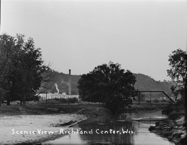 View over river with a small bridge in front of the Carnation Factory with tree-covered hills in the background. Dwellings and fields are in the far right background.