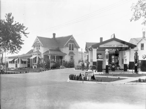 View from street of the Standard Oil filling station with Red Crown gasoline pumps. Two attendants are standing near an entrance with a sign for a postal telegraph office. On the left a woman and two men are standing on the porch of a large house, and a dog is sitting by the stoop. There is decorative running trim along the gables of the house and along the top of the roof, and striped awnings over many of the windows.