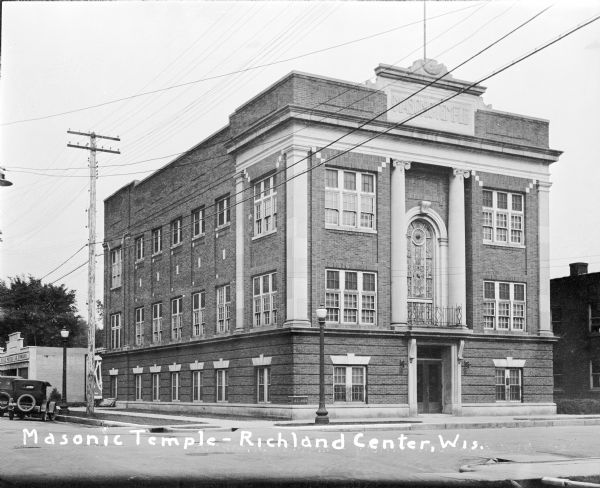 The Masonic Temple on a street corner with lampposts. A large stained glass window and columns above the entrance. Automobiles parked near a service garage back on the left.