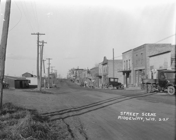 An uphill view of a street in the main business district. The right side is lined with buildings. A drug store, a chiropractor and signs for tires and gas. A truck parked in front of a Mobil Oil vendor.