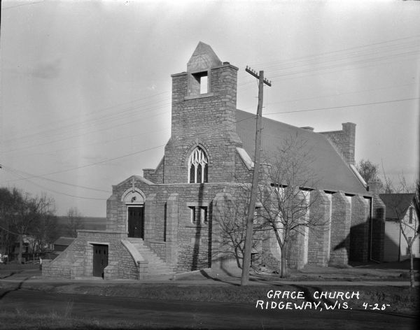 View across road of three-quarter view from front of Grace Church. A double door entrance at the sidewalk level is flanked by stone steps that lead up to another double door entrance. Stone buttresses are along the side of the stone church. In the background are houses and fields.