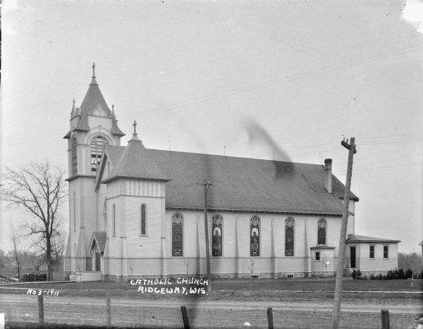 View across road of the Catholic church. Images of saints are in the stained glass windows along the side. There is a large belfry near the front entrance. The wood church has wood buttresses along the side and angled buttresses at the corners.