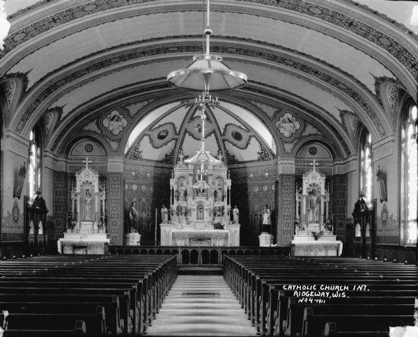 View down center aisle towards the altar. Pews are on both sides of the aisle, and chandeliers hang from the center of the ceiling. There are sculptures of saints flanking the altar which is elaborately carved and has more sculptures in niches. Two smaller altars flank the left and right sides of the church. Paintings of angels holding banners are on the arches as well as the arched ceiling. Stained glass windows are along the sides of the church.