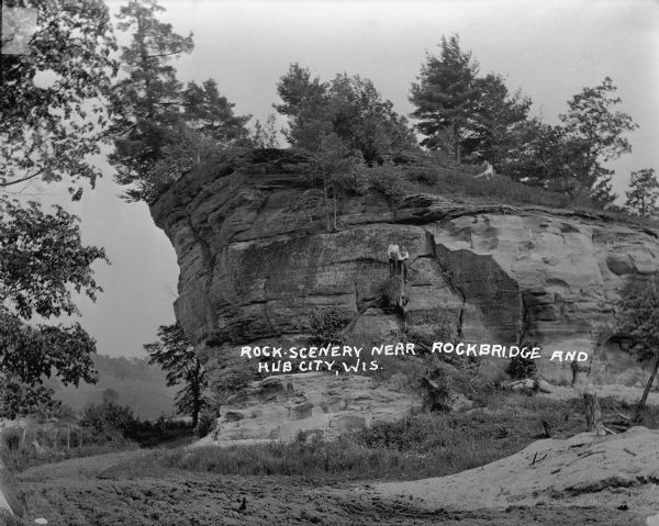 View across dirt road of two men standing on a tree stump halfway up the side of a rocky bluff. Trees grow along the sides and top of the bluff. In the background on the left are fields, trees and rolling hills.