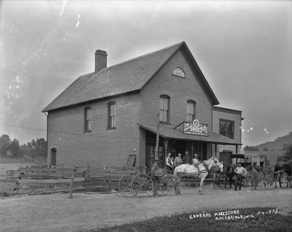 Six men, a young boy and a woman stand outside on the porch and road in front of the General Store. There is a sign for "Star Brand Shoes" posted on the roof of the porch. Above the porch near the peak of the roof in a brick archway is the sign "IOOF 286". Horse-drawn vehicles are parked in the dirt road in front. Wooden fencing is along the left side of the building. In the background are fields, trees, and low hills.