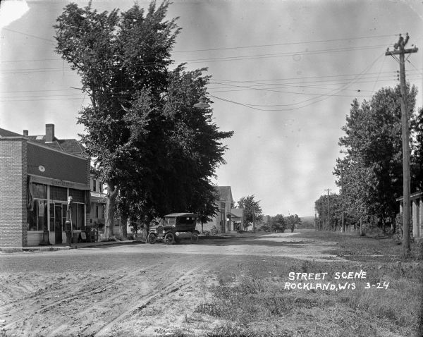 View down unpaved street towards a general store on the left. The sign on the awning in front reads: "Homstad Store." A gas pump, parked car and large trees are in front. Homes and businesses are further down the street along the left.