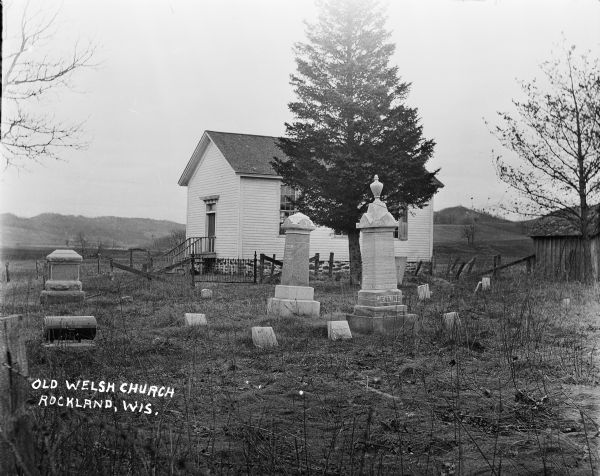 View across graveyard of the Old Welsh Church. The Jenkins family tombstones are in the foreground. Separating the cemetery from the church is a wooden fence with an iron gate. There is a shed on the far right.