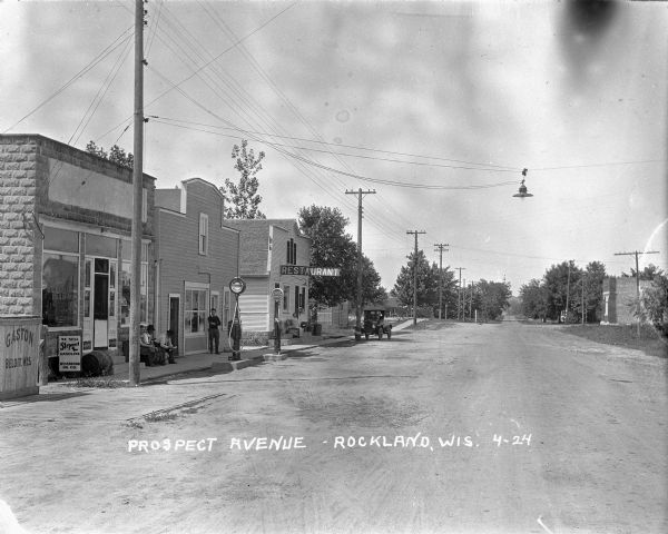 View down Prospect Avenue, with men sitting on a bench by the "Steponet" gas pumps in front of a storefront. A man stands on the sidewalk in front of the building next door. Further down the street a woman sits on the bench in front of a restaurant. A truck is parked along the curb, and a streetlight is suspended above the avenue. There is a small brick building on the right.