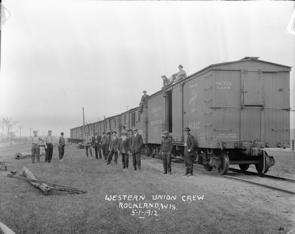 A group of 16 men, some in suits, some in work clothes, posing with railroad cars from the Chicago and Northwestern Railroad.