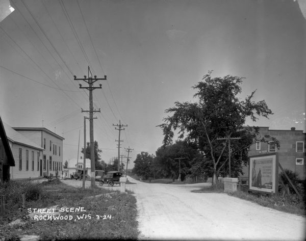 View down unpaved street of a large store on the left with three cars parked in front, near an ice cream signboard. There is a house with a balcony in the background. On the right side of the street is a large, two-story building. In the foreground is a billboard advertising Camel cigarettes, with a stone marker nearby identifying the road as 16 W.
