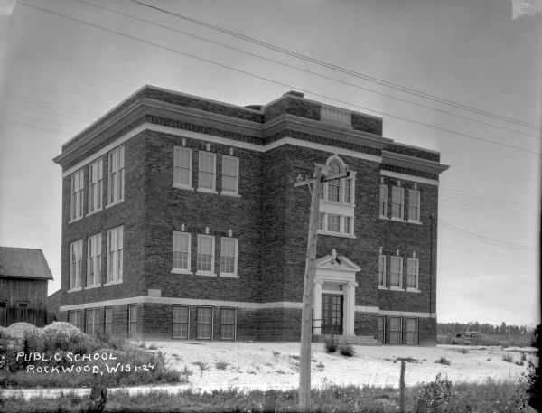 View of the front of the public school, a three-story brick building. Wooden buildings are in the background on the left. In the foreground is a barbed wire fence.