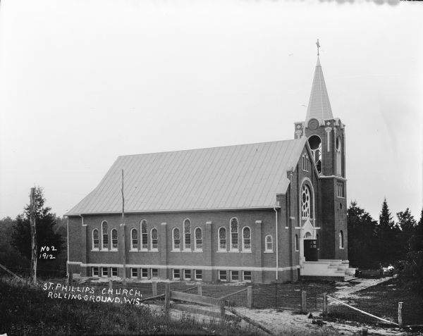 View from hill of the left side of St. Phillips Church, a large brick building with twelve arched stained glass windows, as well as twelve windows along the foundation for the basement. There is a large rose window above the front entrance, and a belfry along the right side. Fencing surrounds the grounds. Near the entrance one man is sitting on a bench, with another man standing nearby.