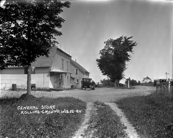 View down path towards a general store with a gas pump on the porch near a sign for Red Crown Gasoline. Two men are working on a car parked in front. Behind them is a large building with a bull in a pasture. Two cars are parked in the road  on the right. In the far background is a house with a windmill.