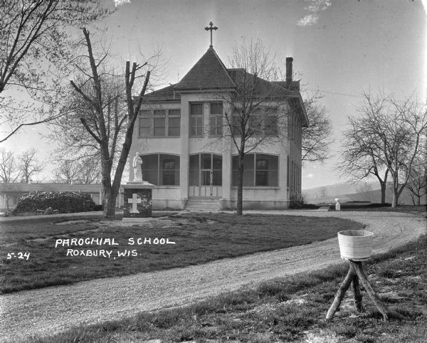 The parochial school with a statue of St. Dominic in the yard in front. There is across on the roof above the entrance. There is a large stack of firewood on the left. A bucket supported by tree limbs is and a near the path in the foreground, and near the sidewalk at the side of the building is a baptismal font.