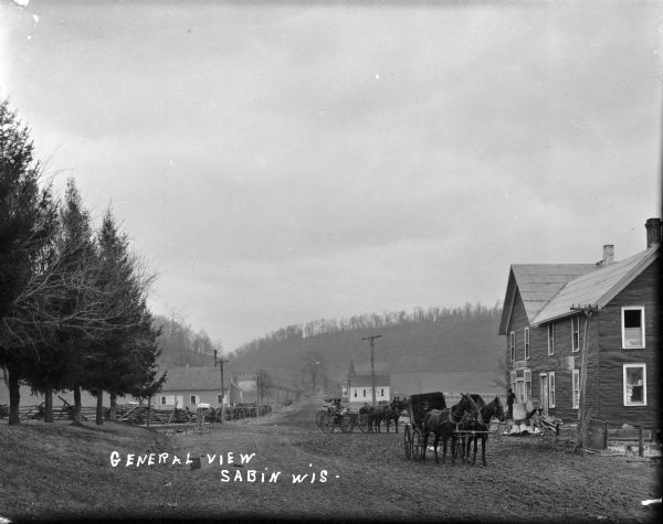 View down unpaved road towards the General Store on the right, with horses and buggies and their drivers assembled out front. A man is standing on the front stoop. The church is behind the buggies further down the road. Two mailboxes stand on the side of the road in front of a split-rail fence, a pasture and more buildings on the left. Hills are in the background.