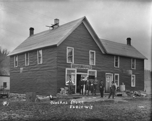 View from unpaved road of the front of the two-story general store with a group of six men standing on the stoop. One man, perhaps the proprietor, wears striped overalls, a bow tie, and leather cuffs over a button down white shirt. The other five men wears suits, hats and ties. A sign above the door reads: "Bargain Counter - Variety of 5 & 10 cent goods." Lacy curtains are in the windows flanking the entrance. Sacks and other items are piled near the open door. An advertisement for shoes in the window on the near right.