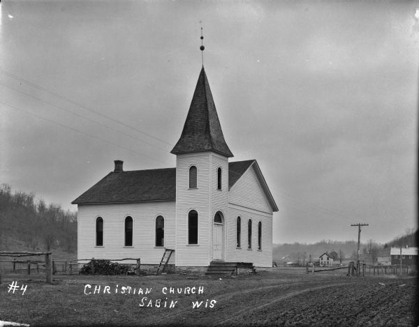 View down unpaved road towards the left side and front of the wood clapboard sided church. There are six arched windows along the side and in the front of the building. More arched windows are on the bell tower over the entrance. The bell tower has a lightning rod on the roof. A stack of firewood is piled along a fence. Houses and farm buildings in the background, and low hills are in the distance.
