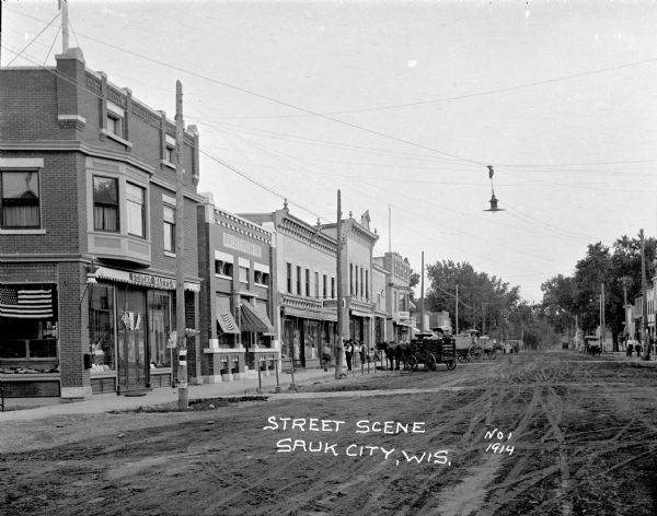 View down unpaved street towards the central business district. There is a drug store on the left decorated with flags, and the Farmers & Citizens Bank and clothing store with a group of men and women standing on the sidewalk in front. Delivery wagons and trucks are parked along the curb. More buildings are on the right, and trees line the street in the background.