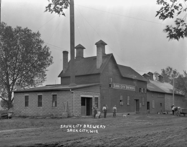 View from unpaved road of workers gathered in front of the brewery building, which has numerous chimneys, and one very tall chimney pipe. One of the men is holding a young child. Two other men stand next to a boy and two dogs. One man leans out a window of the building. A horse and wagon are parked in the background.