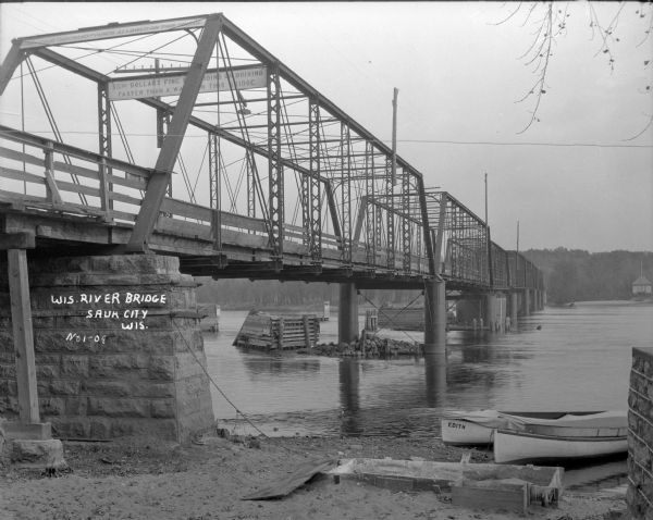 A view from shoreline below a bridge that crosses the Wisconsin River. There are two signs on the upper level of the bridge. One is for weight limit and the other is a warning regarding speed. Two canoes are pulled up on the bank in the foreground.