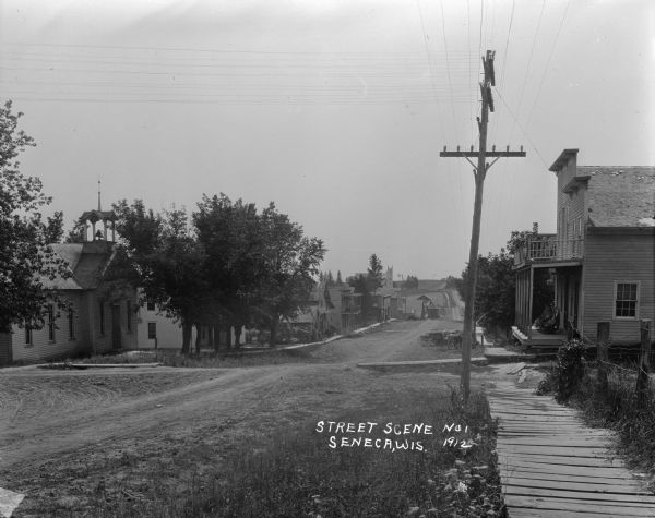 View down unpaved street with a school building on the left on the opposite side of the street. A board sidewalk leads to a two-story dwelling on the right, which has two rocking chairs on the porch with blankets draped over them. In the distance on the left there's a church and a windmill. Between the school and the church are commercial buildings with a man standing in front of one. The right side is tree lined and there are horses and wagons parked in the street.