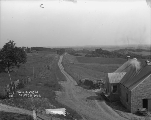 A scenic view over rolling hills with a road leading to the horizon. There are trees, fields and farm buildings on the left. In the foreground on the right is a brick building with milk cans on the porch and a stack of fuelwood in the yard.
