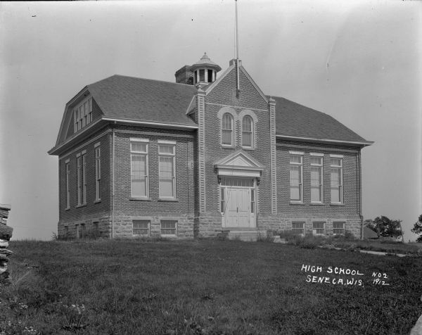 Exterior view of high school, which is a brick building with a ground floor, first floor and attic. A flag pole is above the entrance which has double doors with a triangular pediment, transom windows, two arched windows above it on the first floor. Part of a flagpole extends from the front of the building. Wildflowers are along the front path. Tall windows in the front and side. On the far left is a partial view of a stack of fuelwood.