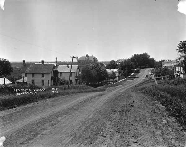 View down an unpaved street on a rolling hill. Wood framed houses and trees are on the left. The high school is in the background.