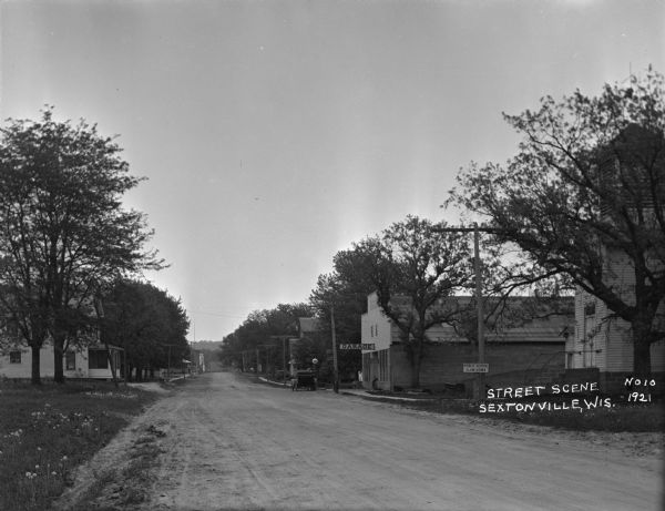 A tree-lined street scene with a house with dandelions in the yard on the left. Two children are playing on the sidewalk in front. On the right is a school with a sign that says: "Public School Slow Down". Next door to the school is a garage with gas pumps. Further down the street is a meat market partially obscured by trees. A man is standing on the curb behind his car near a gas pump.