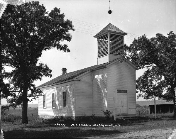 Three-quarter view of the front and side of the M.E. Church, which is a wood frame building. Above the entrance is a bell tower with a weather vane. Near the steps to the front entrance are two posts with a curved wire, presumably for holding lanterns. Two large trees are on either side and a stable is in the background on the right near fields.