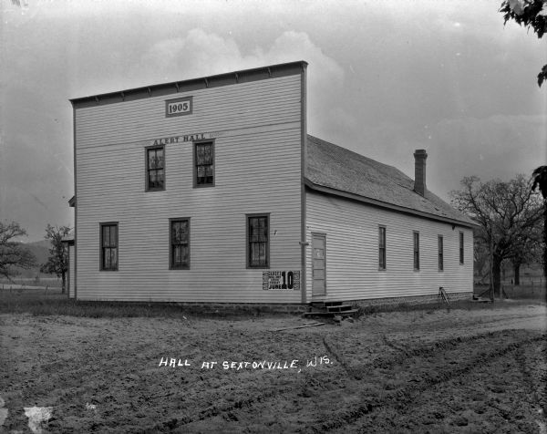 Exterior of Alert Hall. Near the roof line is a sign with the date "1905". Posters are in the windows and a broadside is posted outside underneath a window announcing an upcoming show of "Colored Musical Comedy". Trees, hills and fields are in the background.